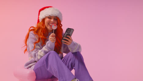 Studio-Shot-Of-Young-Gen-Z-Woman-Wearing-Christmas-Santa-Hat-Eating-Candy-Lollipop-Looking-At-Mobile-Phone-3